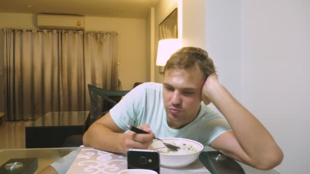 Een moe man zitten aan de tafel in zijn woonkamer, eet diner en via de telefoon. — Stockvideo