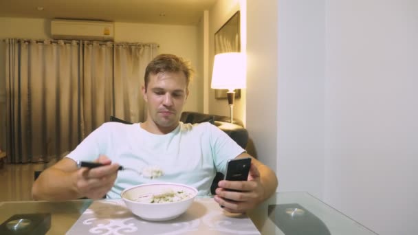 Втомлений чоловік сидить за столом у своїй вітальні, обідає і користується телефоном . — стокове відео