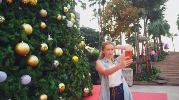 快乐的女人在圣诞树和棕榈树的背景下, 在一个热带城市。新年的概念是到温暖的国家旅行。使用电话 — 图库视频影像