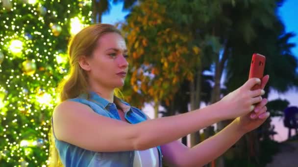 Mujer feliz en el fondo del árbol de Navidad y palmeras en una ciudad tropical. El concepto de Año Nuevo viaja a países cálidos. usando el teléfono — Vídeos de Stock