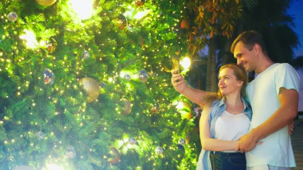 Feliz pareja hombre y mujer haciendo selfie en su teléfono, mientras que de pie contra el fondo de un árbol de Navidad en una ciudad tropical. El concepto de Año Nuevo viaja a países cálidos . — Vídeo de stock