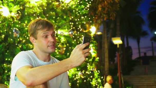 Uomo felice sullo sfondo dell'albero di Natale e delle palme in una città tropicale. Il concetto di Capodanno viaggia in paesi caldi. utilizzando il telefono — Video Stock