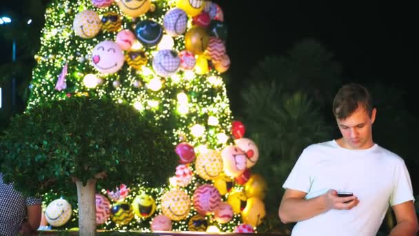 Счастливый человек на фоне елки и пальм в тропическом городе. Концепция Нового года путешествовать в теплые страны. с помощью телефона — стоковое видео