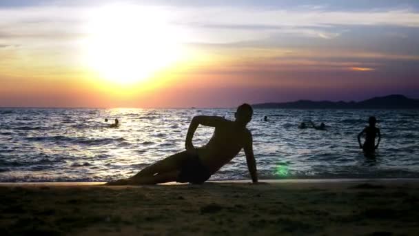 Giocoso bel ragazzo in pantaloncini che fa fitness, in riva al mare sullo sfondo di un meraviglioso tramonto — Video Stock