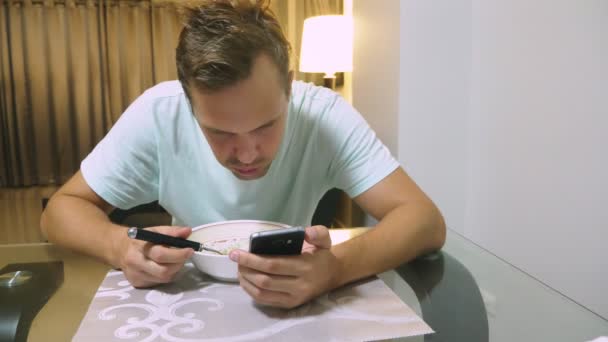 Un hombre cansado está sentado en la mesa de su salón, cenando y usando el teléfono. . — Vídeos de Stock