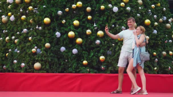 Gelukkige paar man en vrouw selfie maken op hun telefoon terwijl staande tegen de achtergrond van een kerstboom in een tropische stad. Het concept van het Nieuwjaar reizen naar warme landen. — Stockvideo