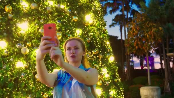 Mujer feliz en el fondo del árbol de Navidad y palmeras en una ciudad tropical. El concepto de Año Nuevo viaja a países cálidos. usando el teléfono — Vídeo de stock