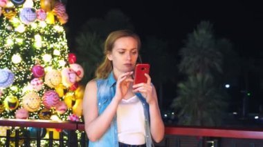 Noel ağacı ve palmiye ağaçları tropikal bir şehirde arka plan üzerinde mutlu bir kadın. Yeni yıl kavramı sıcak ülkelere seyahat. telefonu kullanma