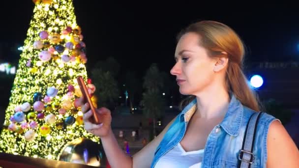 Donna felice sullo sfondo dell'albero di Natale e delle palme in una città tropicale. Il concetto di Capodanno viaggia in paesi caldi. utilizzando il telefono — Video Stock