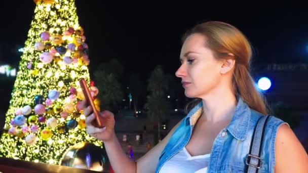 Mujer feliz en el fondo del árbol de Navidad y palmeras en una ciudad tropical. El concepto de Año Nuevo viaja a países cálidos. usando el teléfono — Vídeo de stock