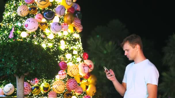 Boldog ember, a háttérben a karácsonyfa és pálmafákkal teli trópusi városban. Új év fogalmának olyan országokba utazhatnak be meleg. a telefon használata — Stock videók