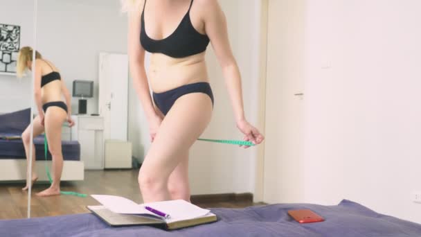 Поняття надмірної ваги і втрати ваги. Жінка вимірює себе вимірювальною стрічкою в спальні, дивлячись на себе в дзеркало, і записує результати в блокнот — стокове відео
