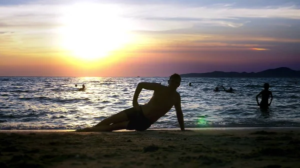 Brincalhão bonito cara em shorts fazendo fitness, na praia contra o pano de fundo de um pôr do sol maravilhoso — Fotografia de Stock