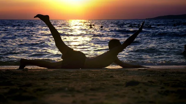 Lekfull stilig kille i shorts gör fitness, på havsstranden mot bakgrund av en underbar solnedgång — Stockfoto