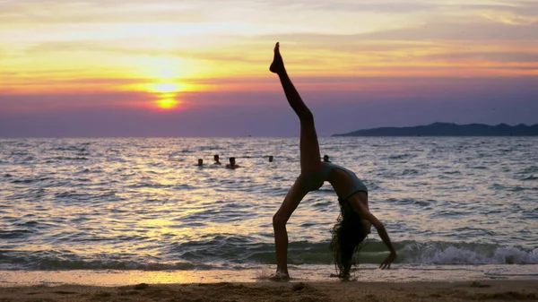 Silueta de una chica al atardecer, contra el fondo del mar, chica delgada piernas graciosa haciendo golpes gimnásticos en la costa del mar — Foto de Stock