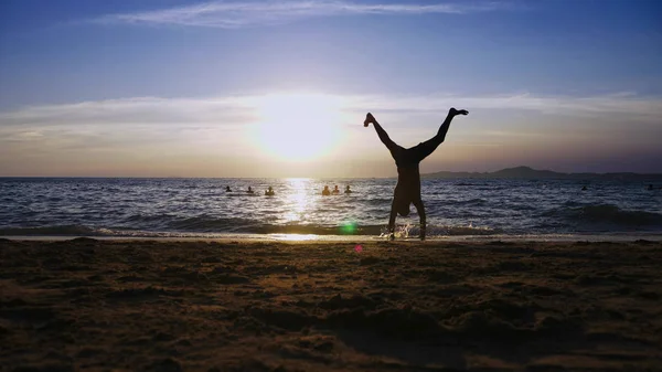 Brincalhão bonito cara em shorts fazendo fitness, na praia contra o pano de fundo de um pôr do sol maravilhoso — Fotografia de Stock