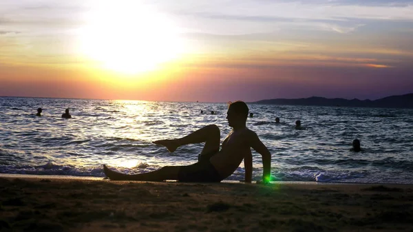 Chico guapo juguetón en pantalones cortos haciendo ejercicio, en la orilla del mar contra el telón de fondo de una maravillosa puesta de sol — Foto de Stock