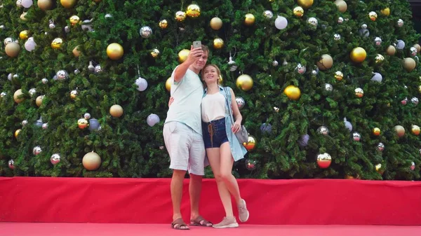 Pasangan bahagia pria dan wanita membuat selfie di telepon mereka sambil berdiri di belakang pohon natal di kota tropis. Konsep Tahun Baru perjalanan ke negara-negara hangat . — Stok Foto