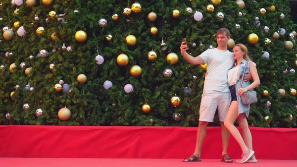 Happy couple man and woman making selfie on their phone while standing against the background of a christmas tree in a tropical city. The concept of New Years travel to warm countries. — Stock Photo, Image