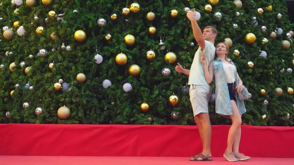 Pasangan bahagia pria dan wanita membuat selfie di telepon mereka sambil berdiri di belakang pohon natal di kota tropis. Konsep Tahun Baru perjalanan ke negara-negara hangat . — Stok Foto