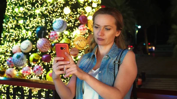 Mujer feliz en el fondo del árbol de Navidad y palmeras en una ciudad tropical. El concepto de Año Nuevo viaja a países cálidos. usando el teléfono —  Fotos de Stock