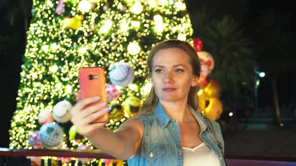Mulher feliz no fundo da árvore de Natal e palmeiras em uma cidade tropical. O conceito de Ano Novo viaja para países quentes. usando o telefone — Fotografia de Stock