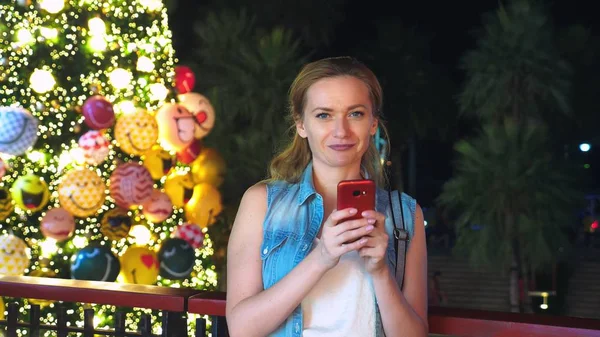 Mulher feliz no fundo da árvore de Natal e palmeiras em uma cidade tropical. O conceito de Ano Novo viaja para países quentes. usando o telefone — Fotografia de Stock