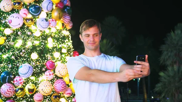 Happy man on the background of the Christmas tree and palm trees in a tropical city. The concept of New Years travel to warm countries. using the phone — Stock Photo, Image