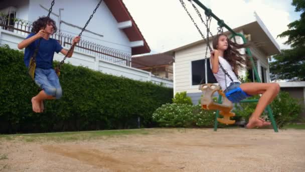 Хлопчик і дівчинка-підліток гойдається на гойдалці з голими ногами на зеленому газоні заднього двору свого будинку — стокове відео