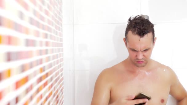 Ein junger Mann studiert die Nachrichten auf seinem Smartphone, während er sich unter der Dusche die Haare wäscht. Konzept zur Internetabhängigkeit — Stockvideo
