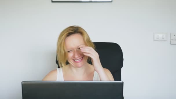 Uma mulher usando seu laptop está sentada à mesa, rindo e conversando. Emoções humanas. Vício em Internet . — Vídeo de Stock