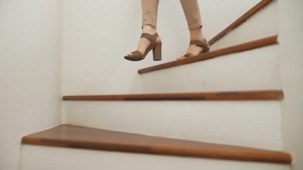 Primer plano, piernas femeninas en pantalones ajustados beige y sandalias de tacón caminando a lo largo de una escalera moderna de madera — Vídeo de stock