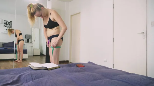 El concepto de sobrepeso y pérdida de peso. Una mujer se mide con una cinta métrica en el dormitorio, mirándose en el espejo, y escribe los resultados en un cuaderno — Foto de Stock