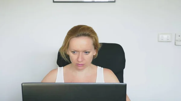 Seorang wanita menggunakan laptopnya, duduk di meja, marah dan kesal, bersumpah. Emosi manusia. Konsep kecanduan internet . — Stok Foto
