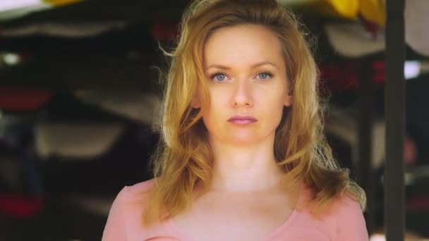 En vacker ung kvinna med blont hår står i ett öppet utrymme på en blåsig dag tittar uppmärksamt på kameran. kopia utrymme — Stockvideo