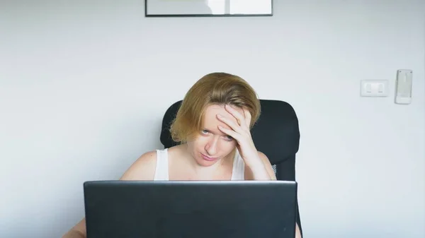 Uma mulher usando seu laptop, sentada à mesa, irritada e irritada, jura. Emoções humanas. conceito de dependência da internet . — Fotografia de Stock