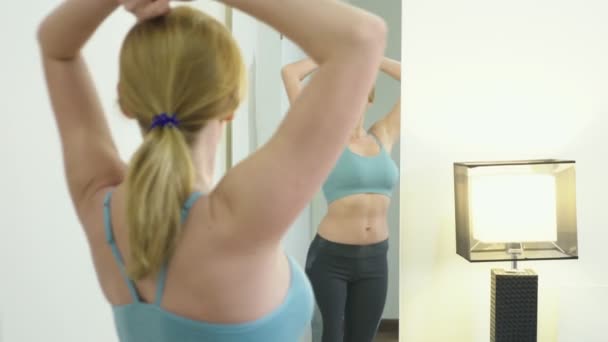 El concepto de exceso de peso y pérdida de peso. Mujer en ropa deportiva mirándose en el espejo en el dormitorio — Vídeos de Stock