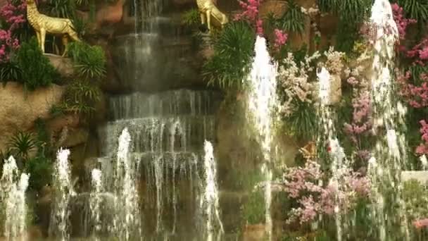 Una gran cascada artificial enmarcada por flores rosadas y hojas verdes . — Vídeos de Stock
