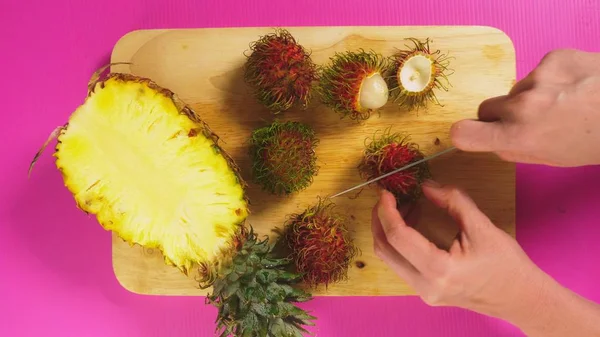 Vista superior, a mão feminina corta frutas com uma faca em uma tábua de madeira, abacaxi e rambutan. O conceito de comida saudável natural . — Fotografia de Stock