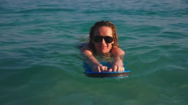 La fille la blonde en maillot de bain noir et des lunettes noires. Un beau modèle avec un corps sexy nage tenant une planche de natation dans de l'eau de mer cristalline . — Video