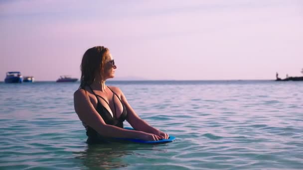 A menina a loira em um maiô preto e óculos pretos. Um modelo bonito com um corpo sexy nada segurando uma placa de natação em água do mar cristalina . — Vídeo de Stock