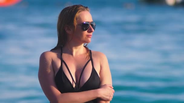 Блондинка в черном купальнике и черных солнечных очках на белом песчаном пляже. Модель с сексуальным телом загорает у моря — стоковое видео