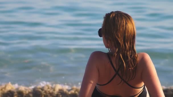 Menina loira em um maiô preto e óculos de sol pretos em uma praia de areia branca. Modelo bonito com corpo sexy banhos de sol pelo mar — Vídeo de Stock