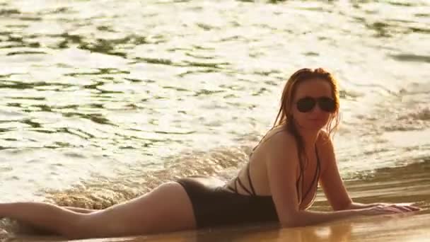 Blonde girl in a black bathing suit and black sunglasses on a white sand beach. Beautiful model with sexy body sunbathes by the sea — Stock Video