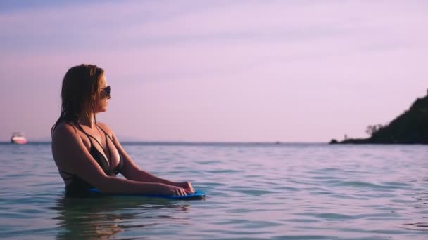 The girl the blonde in a black bathing suit and black glasses. A beautiful model with a sexy body swims holding a swimming board in crystal clear sea water. — Stock Video