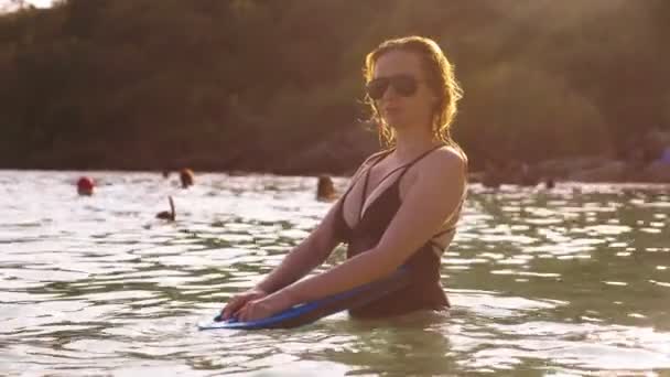 Das Mädchen, die Blondine im schwarzen Badeanzug und schwarzer Brille. ein schönes Model mit einem sexy Körper schwimmt mit einem Schwimmbrett im kristallklaren Meerwasser. — Stockvideo