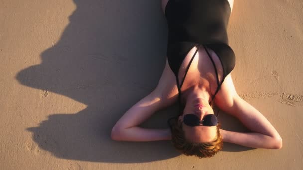 Blondes Mädchen in schwarzem Badeanzug und schwarzer Sonnenbrille an einem weißen Sandstrand. schöne Modell mit sexy Körper Sonnenbäder am Meer — Stockvideo