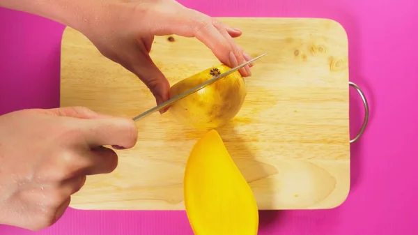 Vista superior, a mão feminina corta frutas com uma faca em uma tábua de madeira, manga. O conceito de comida saudável natural . — Fotografia de Stock