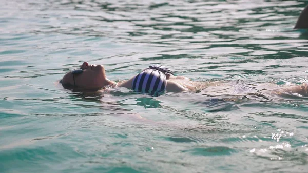 Blond meisje in een badpak en zwarte zonnebril. Mooi model met sexy lichaam zwemt in water van de blauwe zee — Stockfoto
