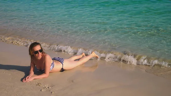 Blond girl in a bathing suit and black sunglasses on a white sand beach by the ocean on vacation. Beautiful model with sexy body swims in blue sea water — Stock Photo, Image